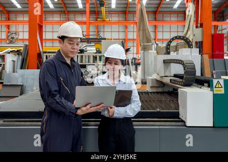 Two worker in a factory examine information on their laptop and tablet computer. They wear safety helmet and work in front of High Precision Fiber Las Stock Photo