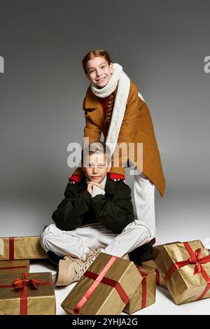 Two cheerful kids dressed warmly pose among beautifully wrapped gifts creating a joyful atmosphere. Stock Photo