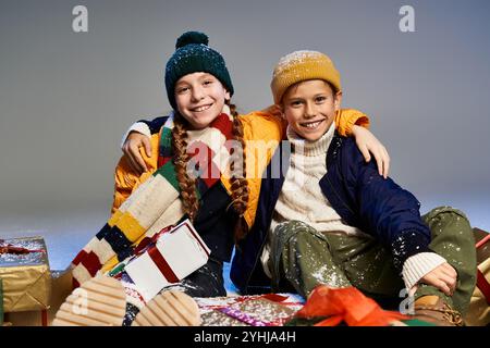 Two cheerful kids dressed warmly share smiles while surrounded by colorful presents and decorations. Stock Photo