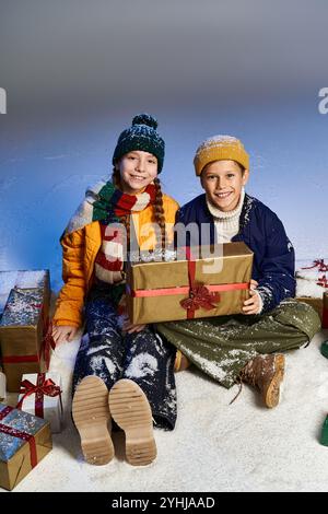 Two happy kids dressed warmly enjoy the winter season, surrounded by festive gifts in snow. Stock Photo