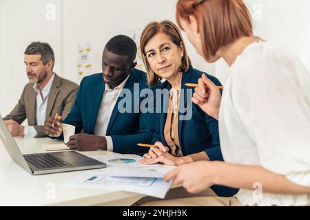 Mature female executive leading diverse business team during financial analysis meeting with multigenerational professionals reviewing data reports Stock Photo