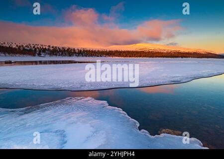 Soft hues of orange and pink paint the sky as the sun sets over a frozen lake. Ice formations edge the water, and distant trees silhouette against the Stock Photo