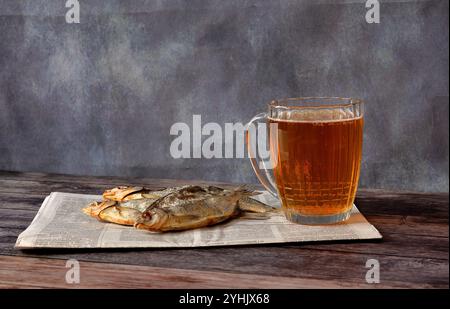 A dark wooden table is covered with newspaper, on which there is a wide mug with light beer and three dried river fish. Close-up. Stock Photo