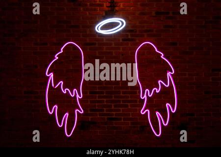 A neon light shaped into one white aureole and two pink angel wings on a brick wall. Stock Photo