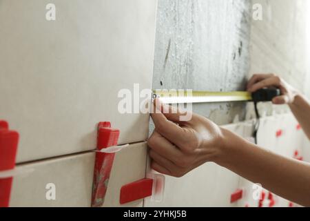 Woman tiling bathroom walls, using tape measure for measuring length. Process of installation of tiles in the bathroom step by step. DIY home improvem Stock Photo