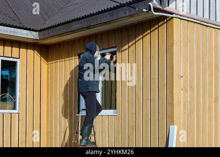 Installing metal window slopes and metal window sills in window opening from facade side, roofing contractor works standing on steel folding ladder. Stock Photo