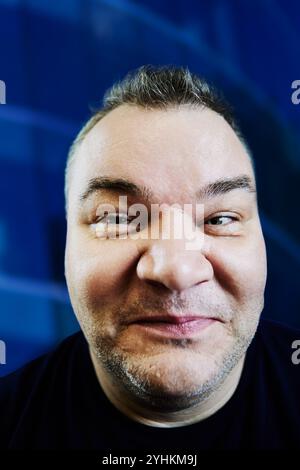 Photographing face with wide-angle lens from close range, 47 year old Caucasian man with an excess weight problem, portrait on blue background. Stock Photo