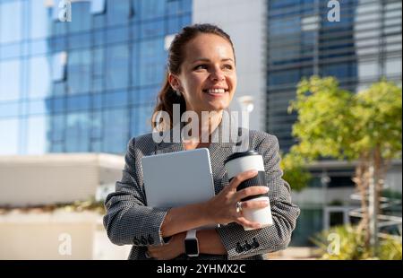 Caucasian young adult businesswoman with digital tablet and travel coffee mug outdoors in downtown area. Stock Photo
