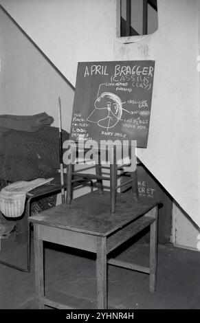 1960, historical, inside a school gym changing room, cricket net and batting pads seen, on a wooden table, a smalll blackboard sitting on a chair. Drawn on it, a funny drawing, a caricature, with the words 'April Bragger' and then in brackets the original name (Cassius Clay) of the famous American heavyweight boxer, Muhammad Ali, who changed his name in 1964. Ali was known for his activism and outspokeness, his boasting, as well as his boxing prowess. The idea behind his 'trash talk' loud vocal style was promotional, a higher profile would attract more paying customers to see his fights. Stock Photo