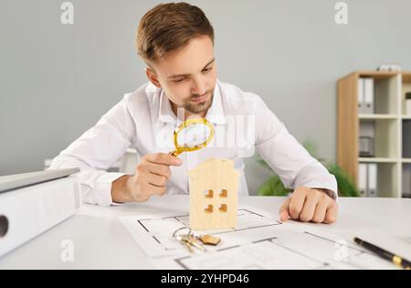 Young man using magnifier to examine wooden house on architecture construction blueprint at desk. Stock Photo