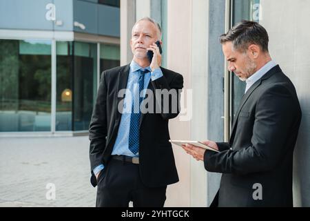 Two professional and successful businessmen standing outside modern office building while one making important phone call and his partner checking information and emails on a tablet during workday. High quality photo Stock Photo