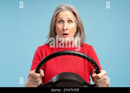 Portrait of sad, fear senior woman holding steering wheel looking at camera standing isolated on blue background. Vehicle, transportation concept Stock Photo