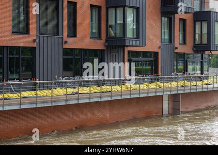 Flood protection measures with yellow sandbags and plastic sheeting safeguard a modern brick building along the riverside. Stock Photo