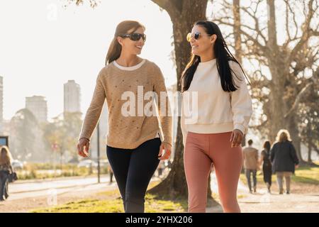 Two women jog side by side in a sunlit park, smiling as they embrace an active lifestyle. Surrounded by lush trees, they enjoy their outdoor exercise Stock Photo