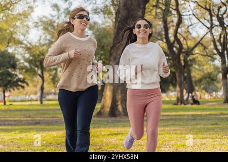 Two women jog side by side in a sunny park, smiling and enjoying the fresh air. Dressed casually in sweaters, they embrace a healthy lifestyle, surrou Stock Photo