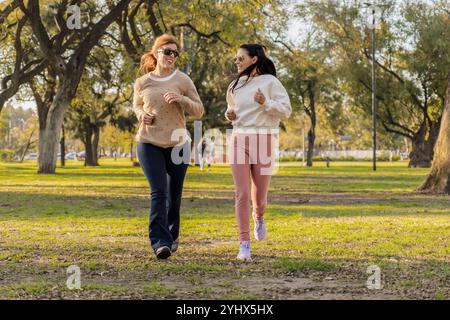 Two women jog side by side in a sunlit park, smiling as they embrace an active lifestyle. Surrounded by lush trees, they enjoy their outdoor exercise Stock Photo