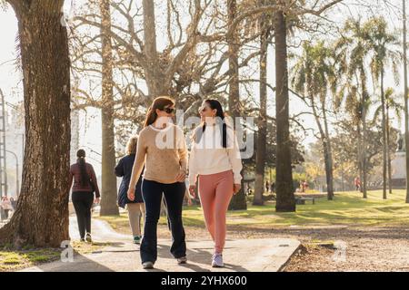 Two women enjoying a walk in the park on a sunny day, engaging in conversation while surrounded by trees. They wear casual outfits, with one in a ligh Stock Photo