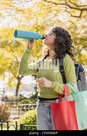 Woman with curly hair and sunglasses drinks from a blue water bottle in a park. She carries reusable bags and wears a green sweater, embodying an eco- Stock Photo