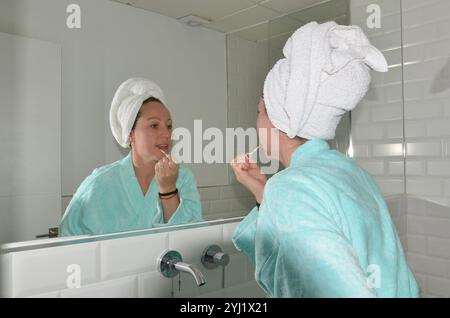 Woman wearing bathrobe and towel applying lip gloss while looking in the mirror in a modern bathroom Stock Photo