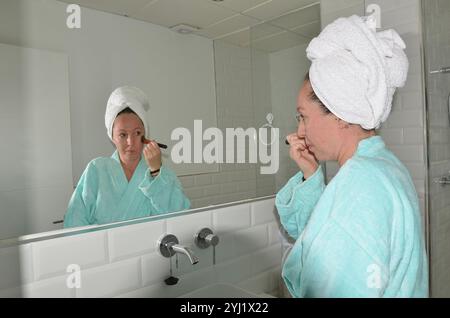Woman applying makeup with brush in bathroom mirror wearing bathrobe and towel turban Stock Photo