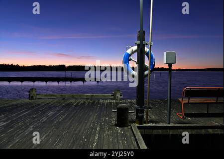 a tranquil lakeside dock scene at sunset or dusk, with a vibrant gradient sky transitioning from orange and pink near the horizon to deep blue overhea Stock Photo