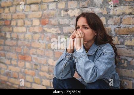 Frustrated Caucasian woman sits next to brick wall. Stock Photo