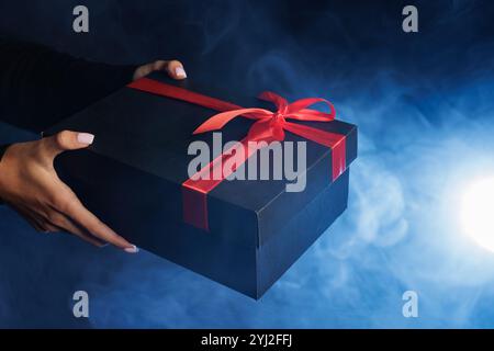 A person holding a black gift box with a red ribbon against a smoky blue background, creating a mysterious and festive atmosphere. Stock Photo