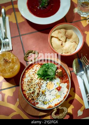 A top-down view of a vibrant Middle Eastern meal featuring shakshuka, beetroot soup, pita bread, and an orange drink on a patterned table. Stock Photo