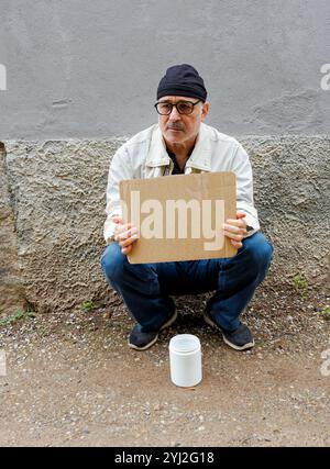 Man squatting against a wall holding a blank cardboard sign with a white container to collect money beside him on the ground, Italy Stock Photo