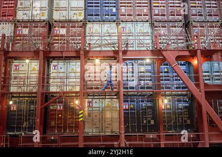 Worker in blue overalls walking along the gangway of a large red cargo ship stacked with colorful containers. Stock Photo