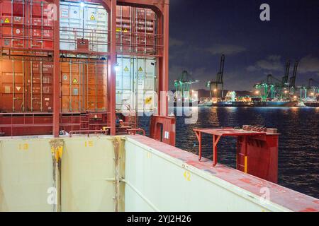 Cargo ship loaded with containers docked at a busy port during nighttime with brightly lit cranes in the background, Singapore Stock Photo