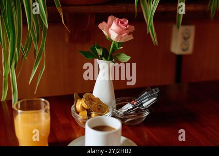 A cozy breakfast setup in a hotel room with a cup of coffee, glass of orange juice, fresh cut rose in a vase, sliced lemon, and pens on a wooden table. Stock Photo