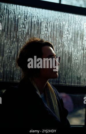 Silhouette of a woman with glasses profiled against a window covered in condensation during golden hour. Stock Photo