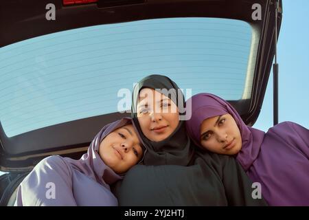 Three women wearing hijabs smiling while sitting in the trunk of a car under a clear sky, Morocco Stock Photo