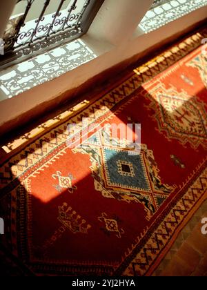 Sunlight streams through an ornate window onto a vibrant red Persian carpet laid on a wooden floor, Morocco Stock Photo