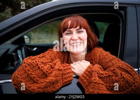 Smiling woman with red hair wearing a chunky knit sweater leaning out of a car window. Stock Photo