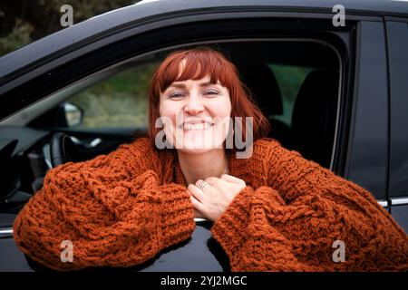 Smiling woman with red hair leaning out from a car window wearing an orange knit sweater. Stock Photo