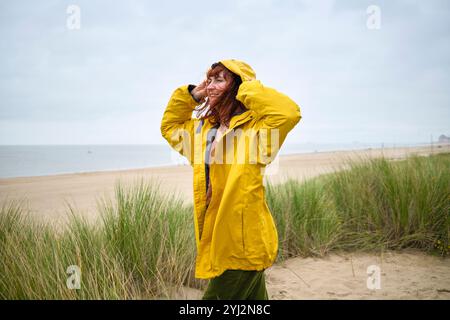 A cheerful woman in a yellow raincoat stands by the beach with grassy dunes, smiling and holding her hair on a breezy day, Belgium Stock Photo