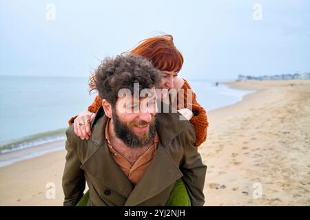 Smiling man with a beard giving a piggyback ride to a happy woman on a cloudy beach day, Belgium Stock Photo