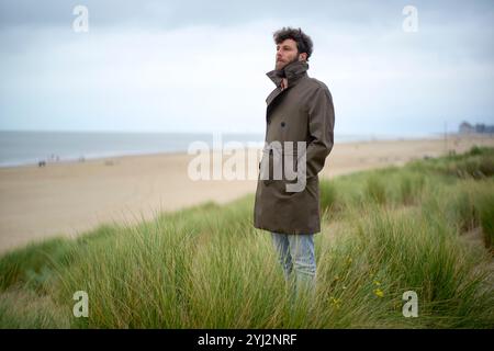 A man in a coat standing thoughtfully in a grassy dune overlooking the beach on an overcast day, Belgium Stock Photo