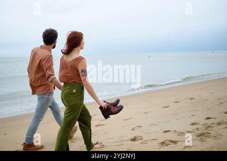 Couple holding hands while walking barefoot along a sandy beach with shoes in hand, with a calm ocean and overcast sky in the background, Belgium Stock Photo