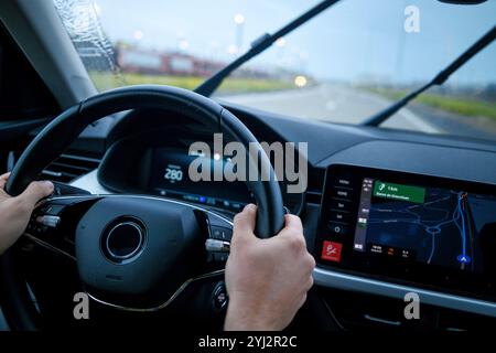 Hands holding the steering wheel of a car with a navigation system on the dashboard displaying a map during a journey. Stock Photo