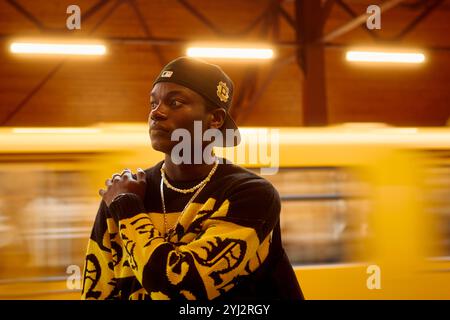 Young man in a black cap and patterned sweater stands pensively against a blurred yellow subway passing by, Berlin, Germany Stock Photo