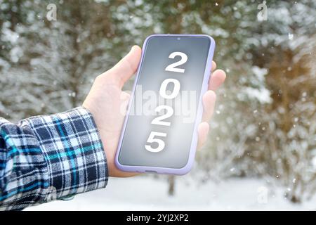 a hand holding a smartphone displaying the number 2025 on the screen, symbolizing the New Year. The background features a winter scene Stock Photo