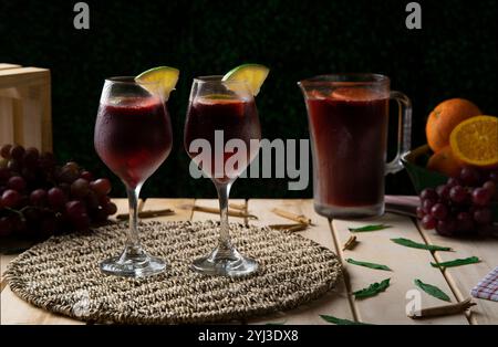 Two glasses of handmade summer wine next to wine jug, oranges and grapes on rustic wooden table against dark defocused background Stock Photo