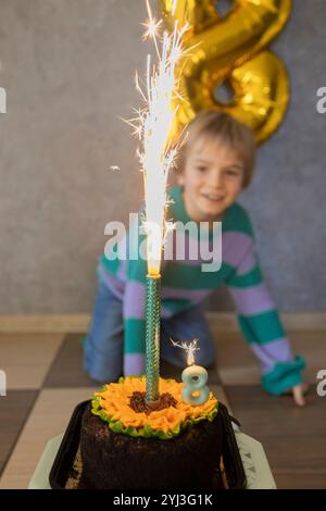 Charming happy blond 8 year old boy celebrating his birthday. The birthday boy looks with delight at the burning candle on the cake. selective focus. Stock Photo