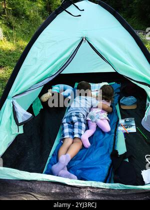 Two children lying inside a tent, enjoying a peaceful camping experience in a lush green setting. The scene conveys a sense of adventure, family bondi Stock Photo