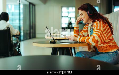 A cheerful high school student engages in text messaging on her smartphone while studying in a contemporary setting, wearing headphones and smiling. Stock Photo