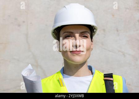 Portrait of a confident female architect wearing a protective vest, smiling and holding blueprints at a construction site Stock Photo