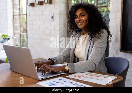 Smiling woman working on laptop at home office with charts on desk Stock Photo